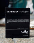 Black detergent sheet box with white writing in front of background of a city sidewalk and people walking.